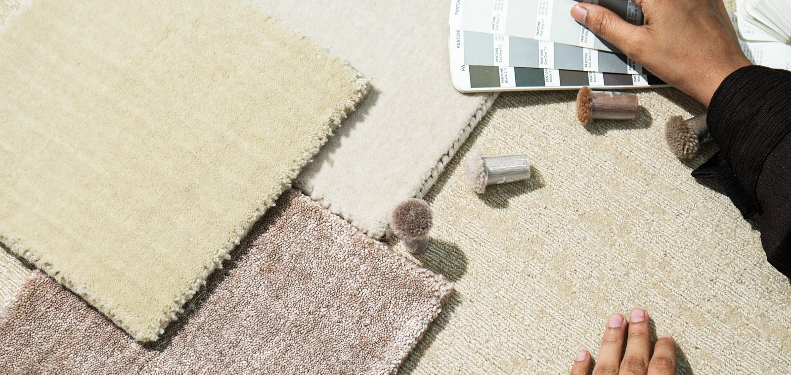 A hand holding a colour swatch fan over rug samples in cream, brown and beige laid out on the floor