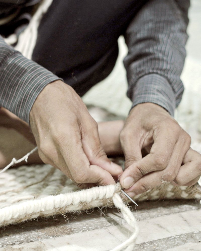 Close-up of two hands binding the edge of a rug with white cord and a needle